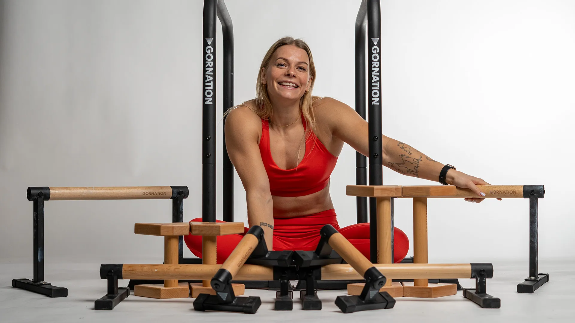 women named Malin Malle sitting with many of different paralletes and dip bars on the ground. Wearing a red sport bra and leggings.