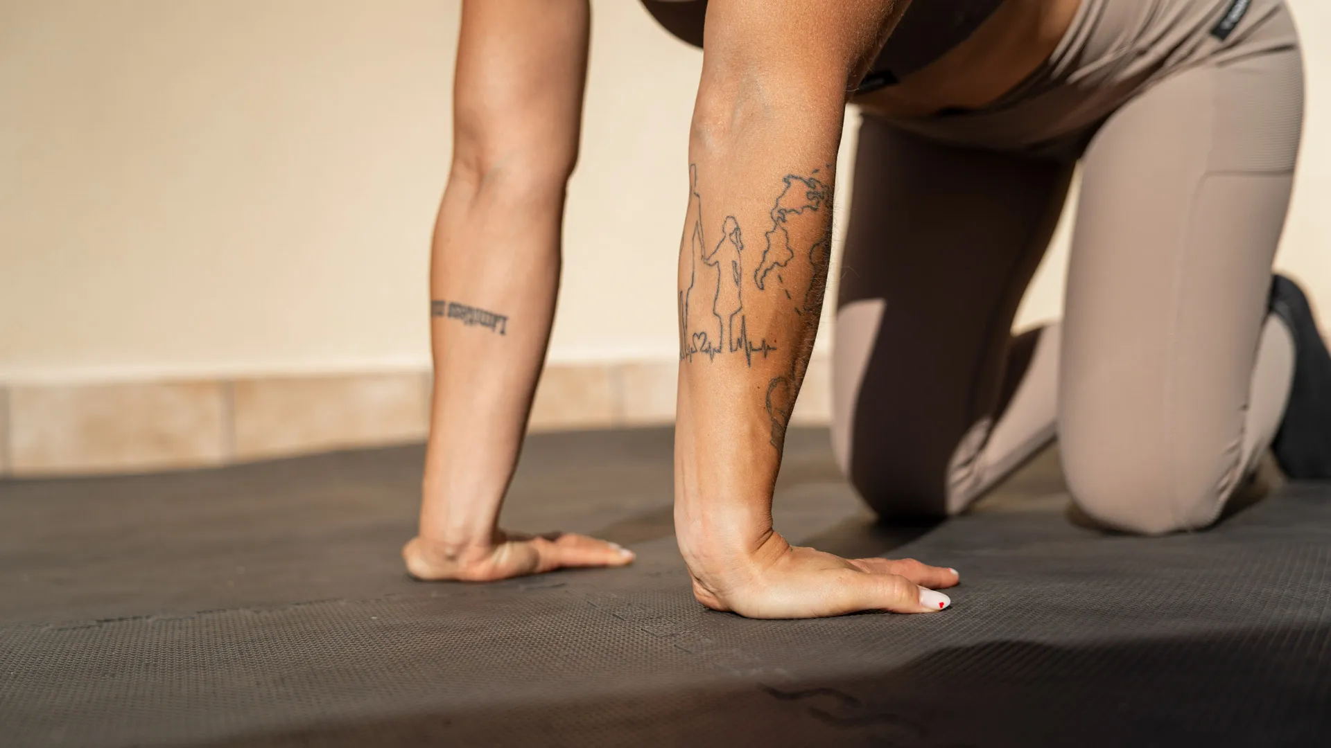 A close-up of a woman's hands and forearms resting on a gym mat in a kneeling position, with visible tattoos, focusing on her form during a workout.