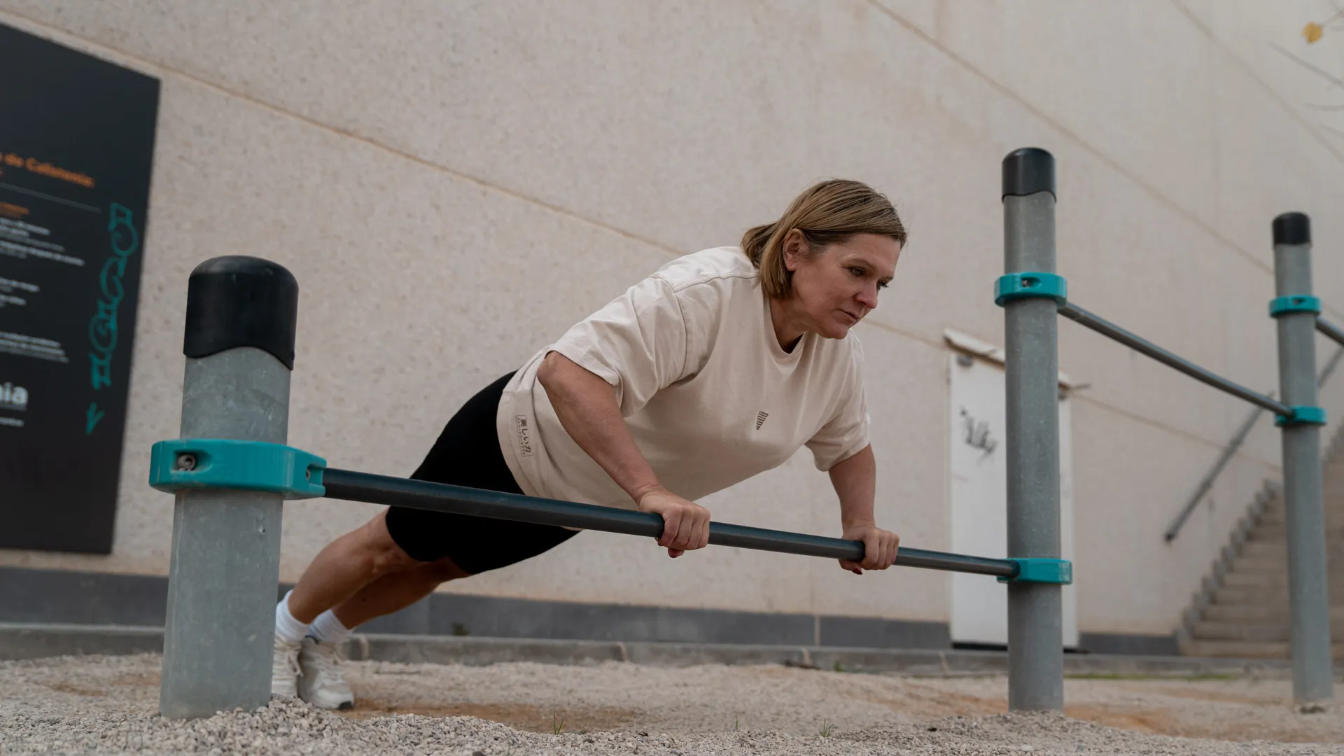Woman performing a push-up on parallel bars outdoors, focused on her form while wearing a white t-shirt and black shorts. The background features a neutral wall and minimalist outdoor calisthenics equipment, highlighting strength and determination in a simple, clean fitness environment. Ideal for calisthenics and strength training enthusiasts.
