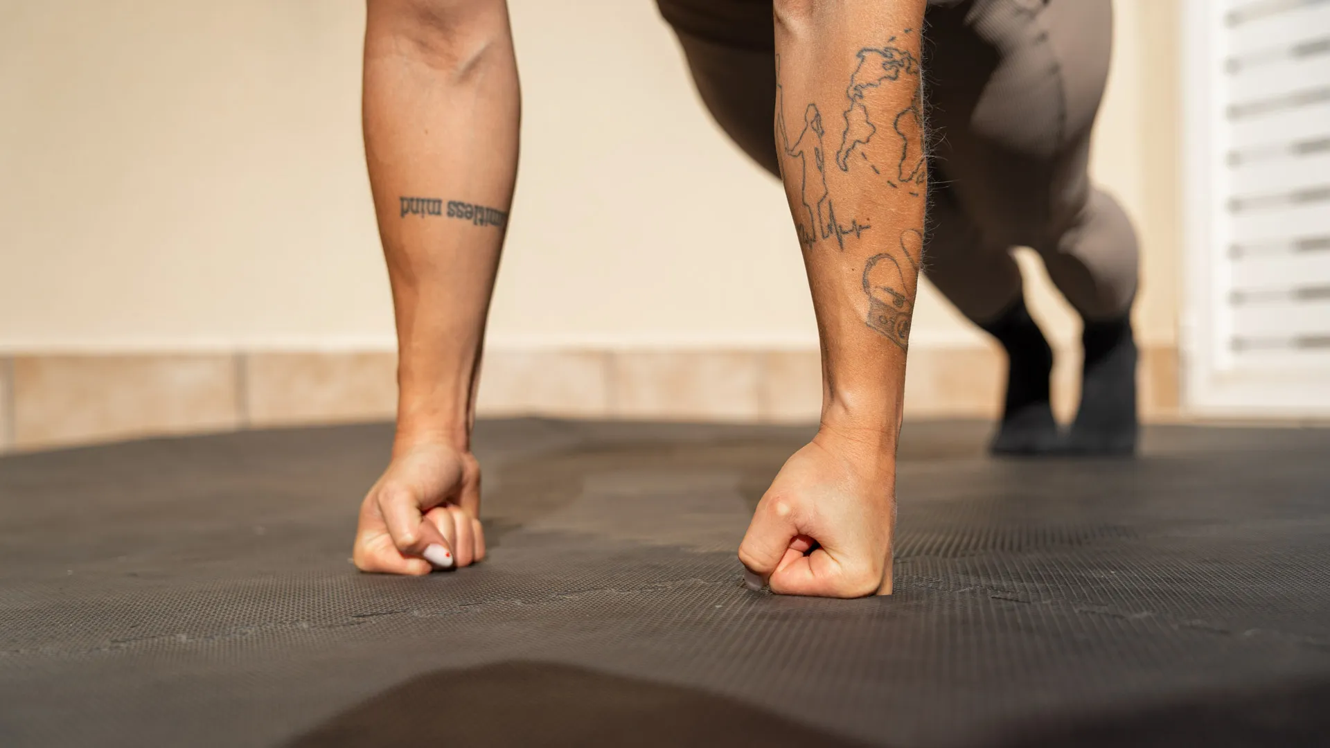 A close-up of a woman's hands in a fist position on a gym mat, demonstrating a push-up or plank position, with tattoos visible on her forearms and a blurred background.