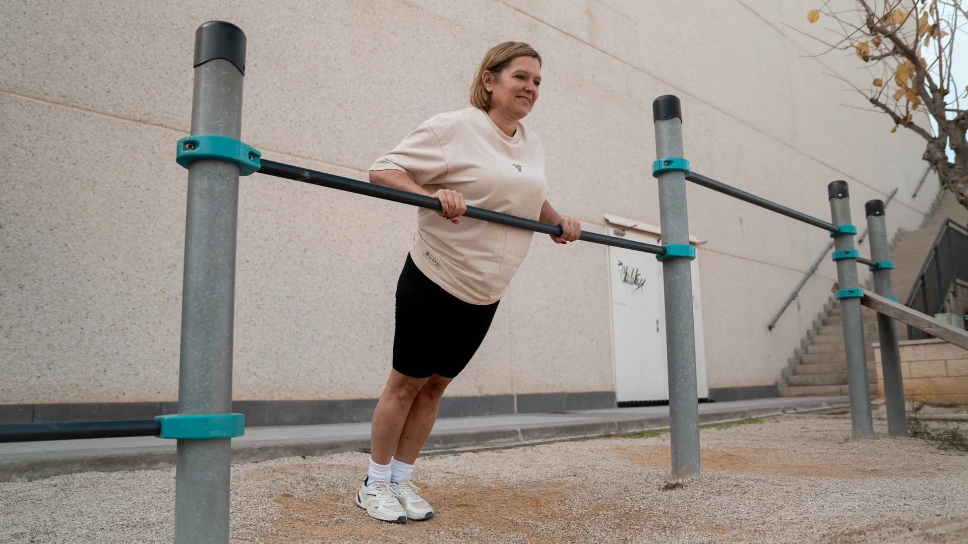 A woman performs an incline push-up on outdoor parallel bars, smiling with a beige wall and stairs in the background.