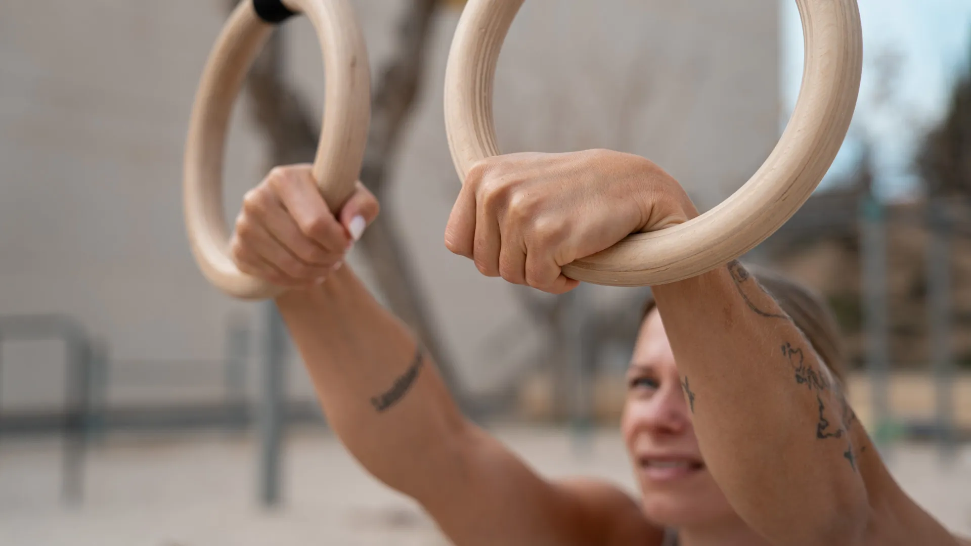 A close-up of a woman's hands gripping wooden gymnastic rings, showcasing her strong hold and form, with a focused expression and a blurred outdoor background.