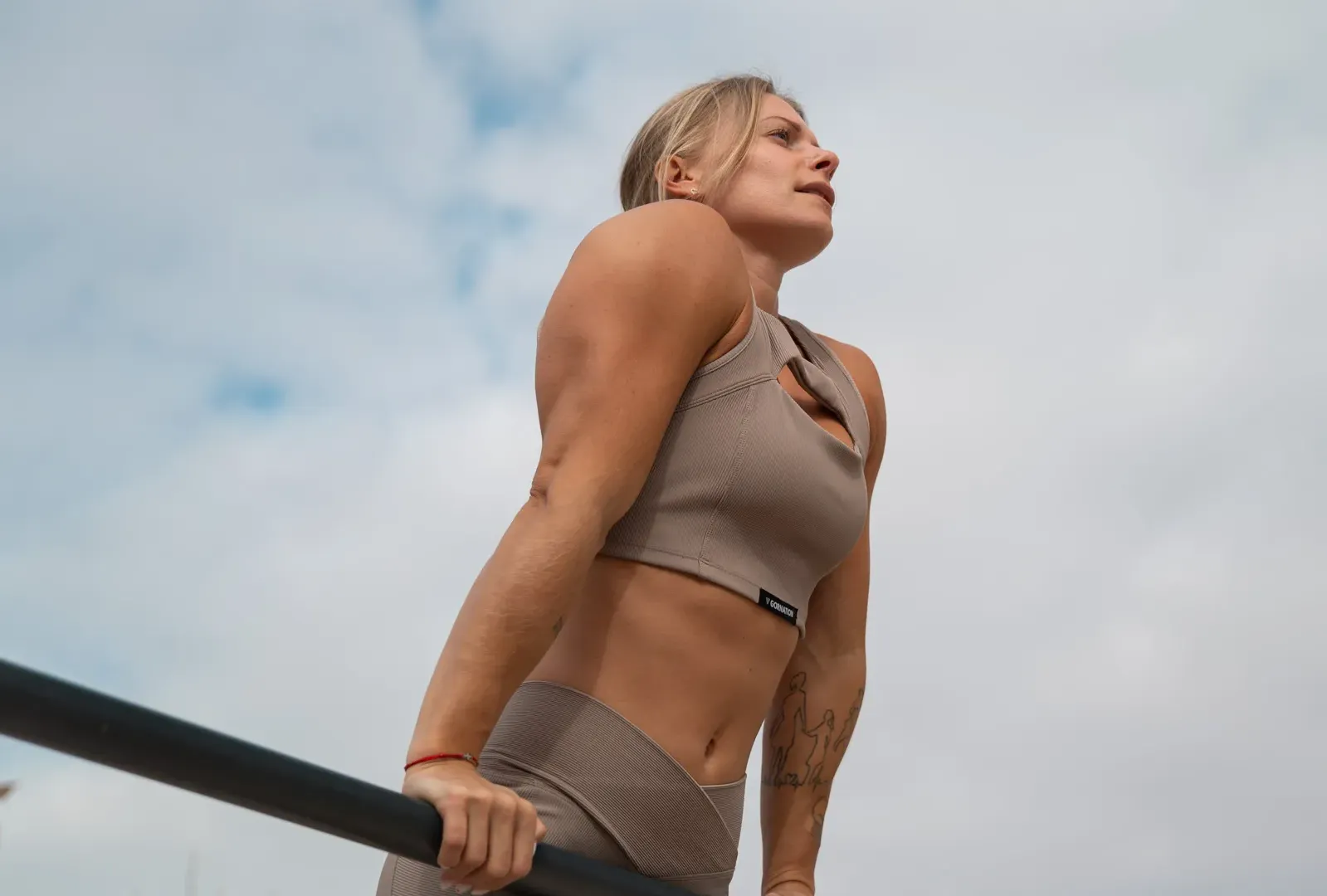 A focused woman in a beige workout set performing a straight bar dip outdoors, highlighting her upper body strength against a cloudy sky.