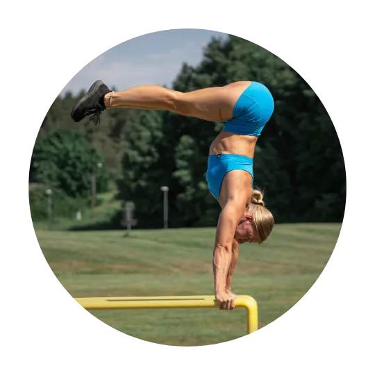 Kvinna som utför en avancerad handstand på parallella stänger utomhus i blå träningskläder.