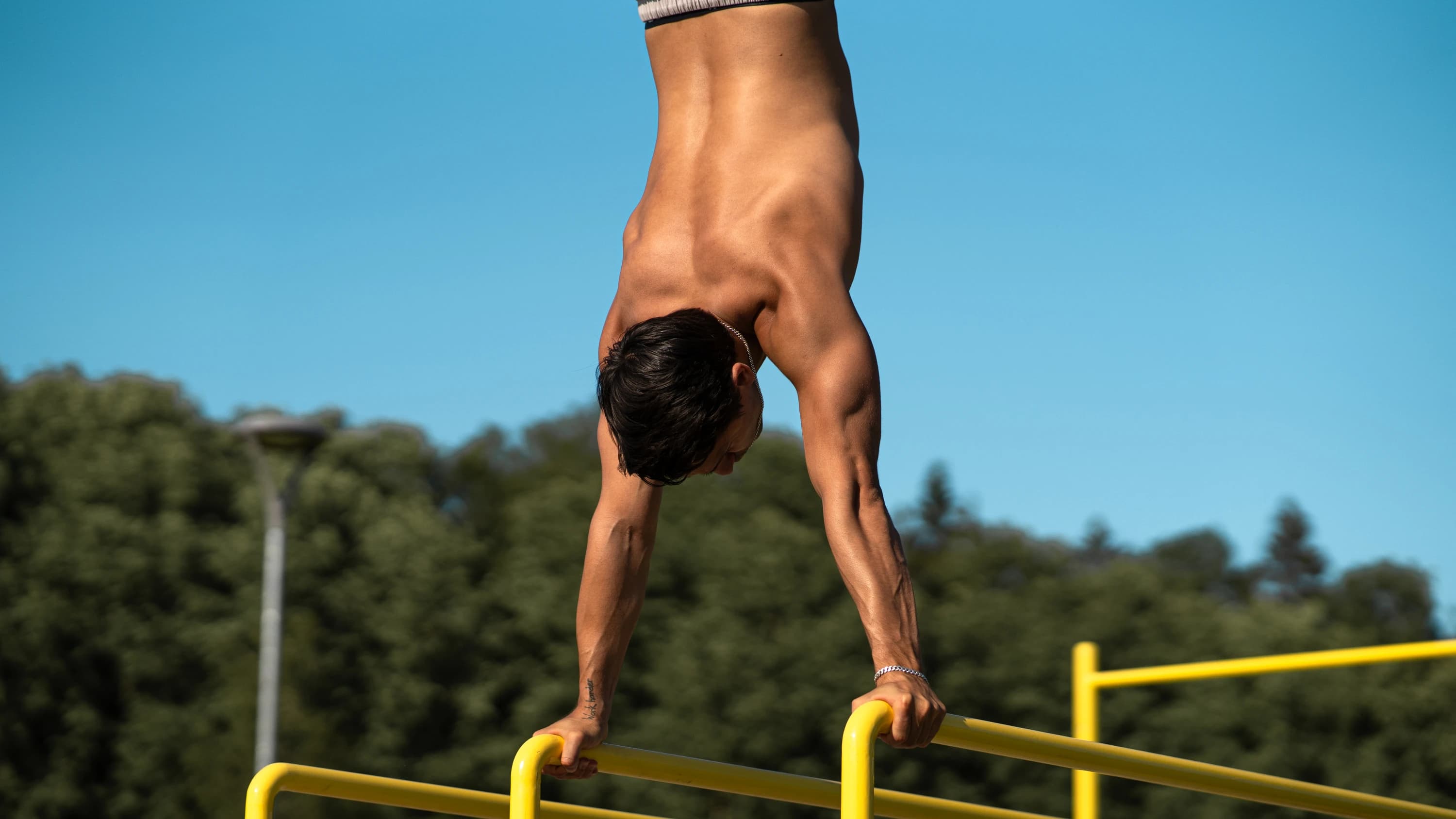 A shirtless man is performing a controlled handstand on yellow parallel bars in an outdoor fitness area. His arms are straight, with hands firmly gripping the bars, and his body is aligned in a vertical position, demonstrating impressive strength and balance.