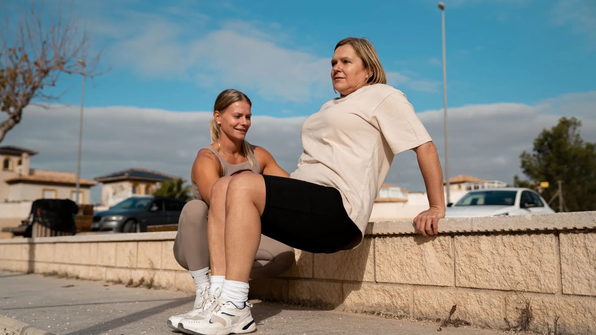 A trainer assists a woman doing tricep dips on a low wall outdoors, with a sunny suburban backdrop.