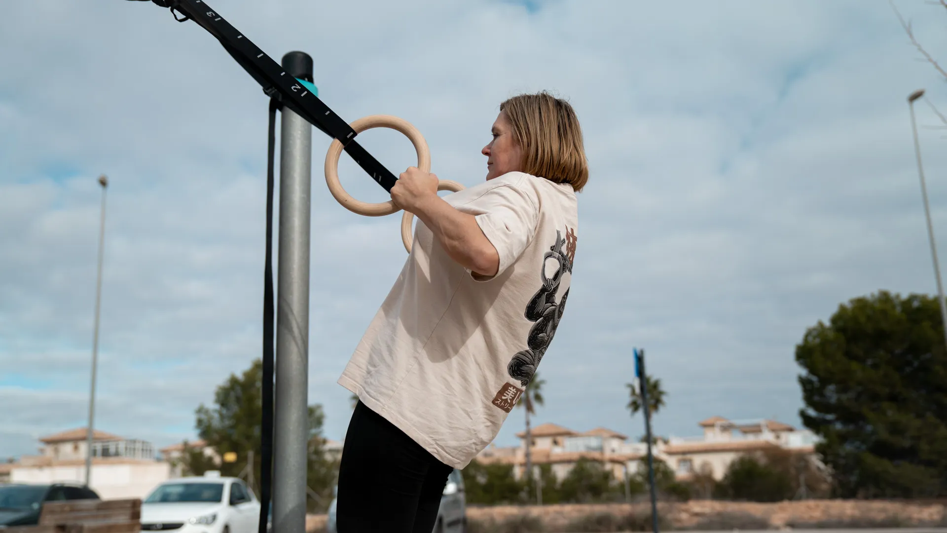 A woman practices ring rows on wooden gymnastic rings outdoors, with a cloudy sky and suburban houses in the background.