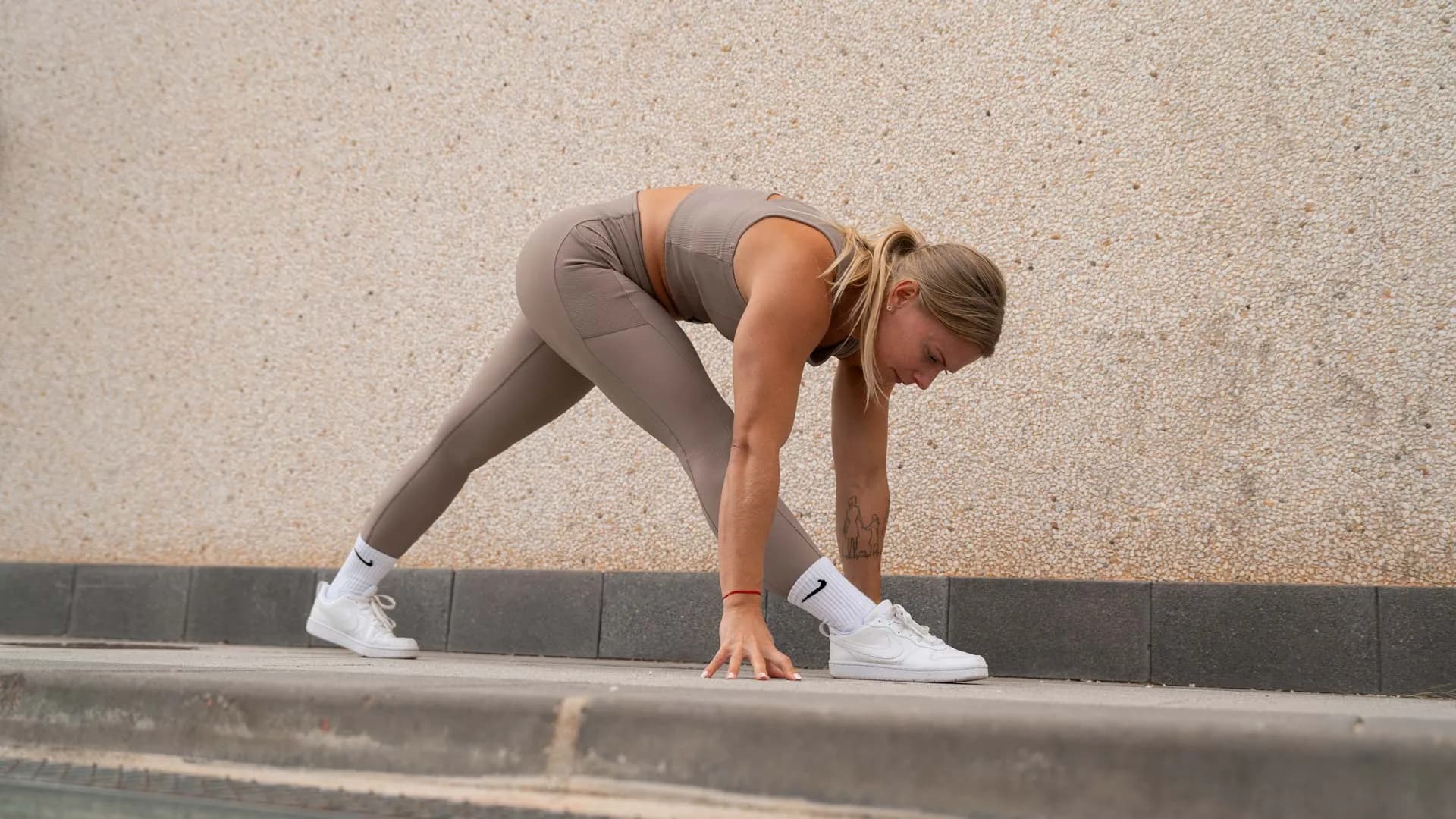 A woman stretches in a forward lunge position, touching the ground with one hand, against a textured beige wall.
