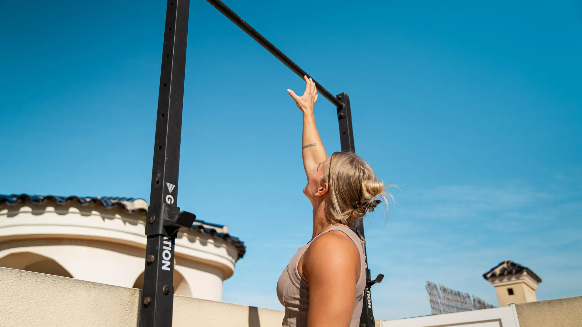 Kvinna sträcker upp mot pull-up-stången i en utomhus-träningsmiljö, i beige träningskläder med klar blå himmel i bakgrunden.