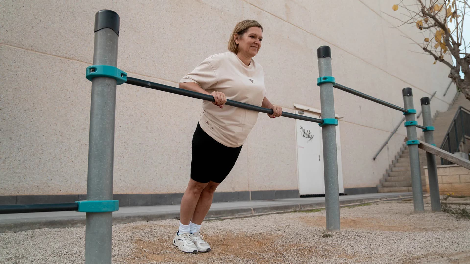 Woman performing an assisted dip exercise on outdoor parallel bars, wearing a white t-shirt and black shorts, showcasing a strong and focused workout routine. The background features a neutral wall, with a minimalistic and clean outdoor fitness environment, ideal for calisthenics and strength training enthusiasts.
