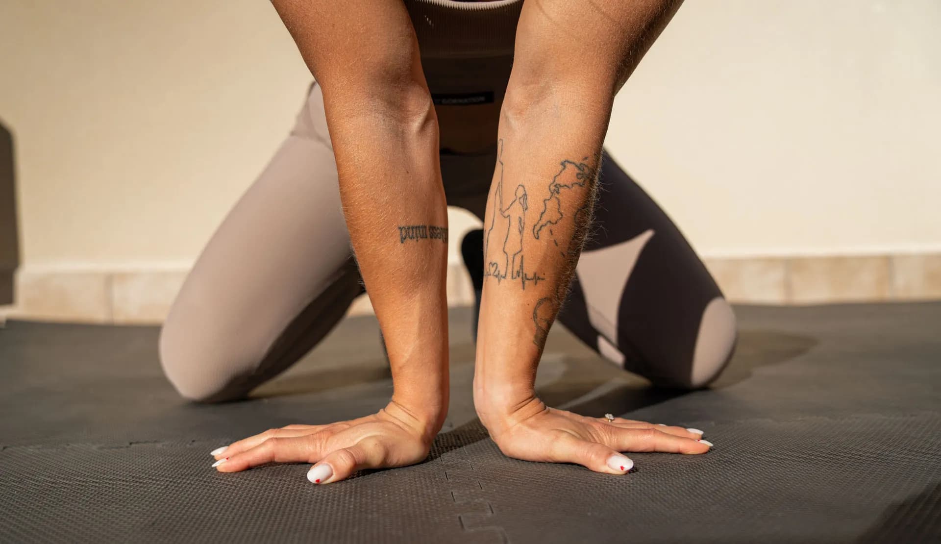 Close-up of a woman's hands and forearms in a wrist stretch position on a workout mat, highlighting her tattoos and flexibility.