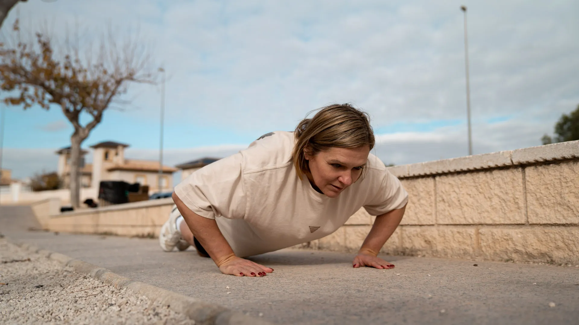 Woman performing a push-up on the ground outdoors, with a focused expression, wearing a beige t-shirt and white sneakers. The background features a minimalist fitness environment with clear skies, trees, and a few residential buildings, showcasing a dedication to bodyweight training and strength exercises.
